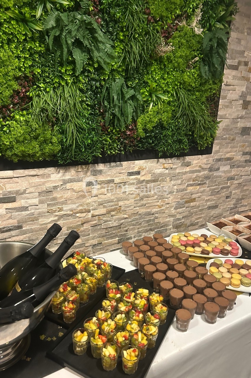 Buffet avec desserts variés, verrines de fruits, macarons et bouteilles de vin, devant un mur végétalisé.