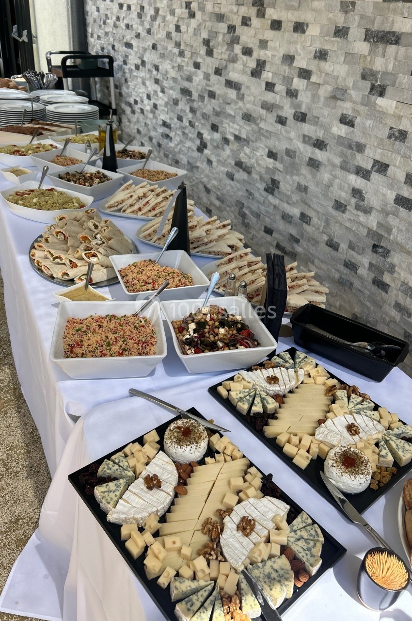 Buffet avec plateaux de fromages, salades variées et sandwichs disposés sur une table blanche en extérieur.