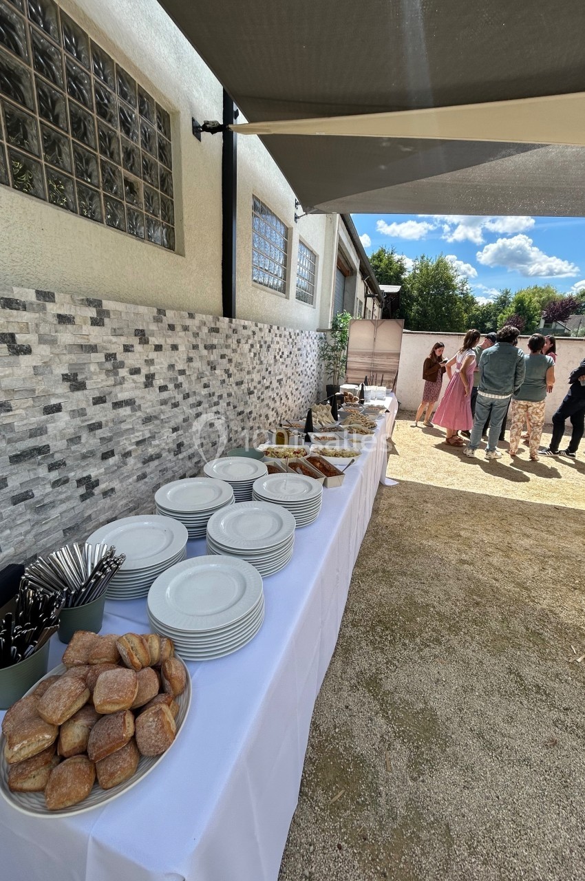 Table dressée en extérieur avec assiettes, couverts, pains et viennoiseries, des invités discutent en arrière-plan.