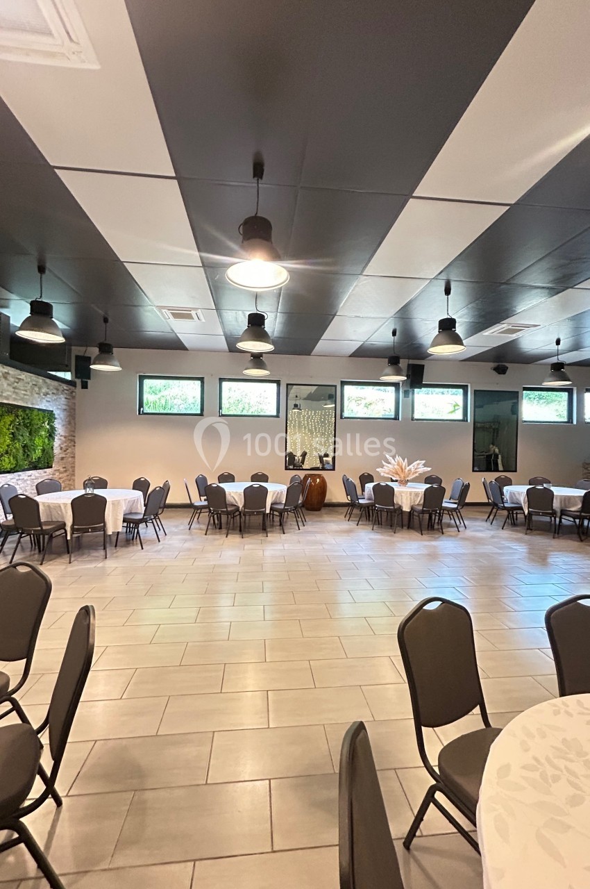 Salle spacieuse avec tables rondes dressées, chaises noires, murs en pierre et végétation décorative.