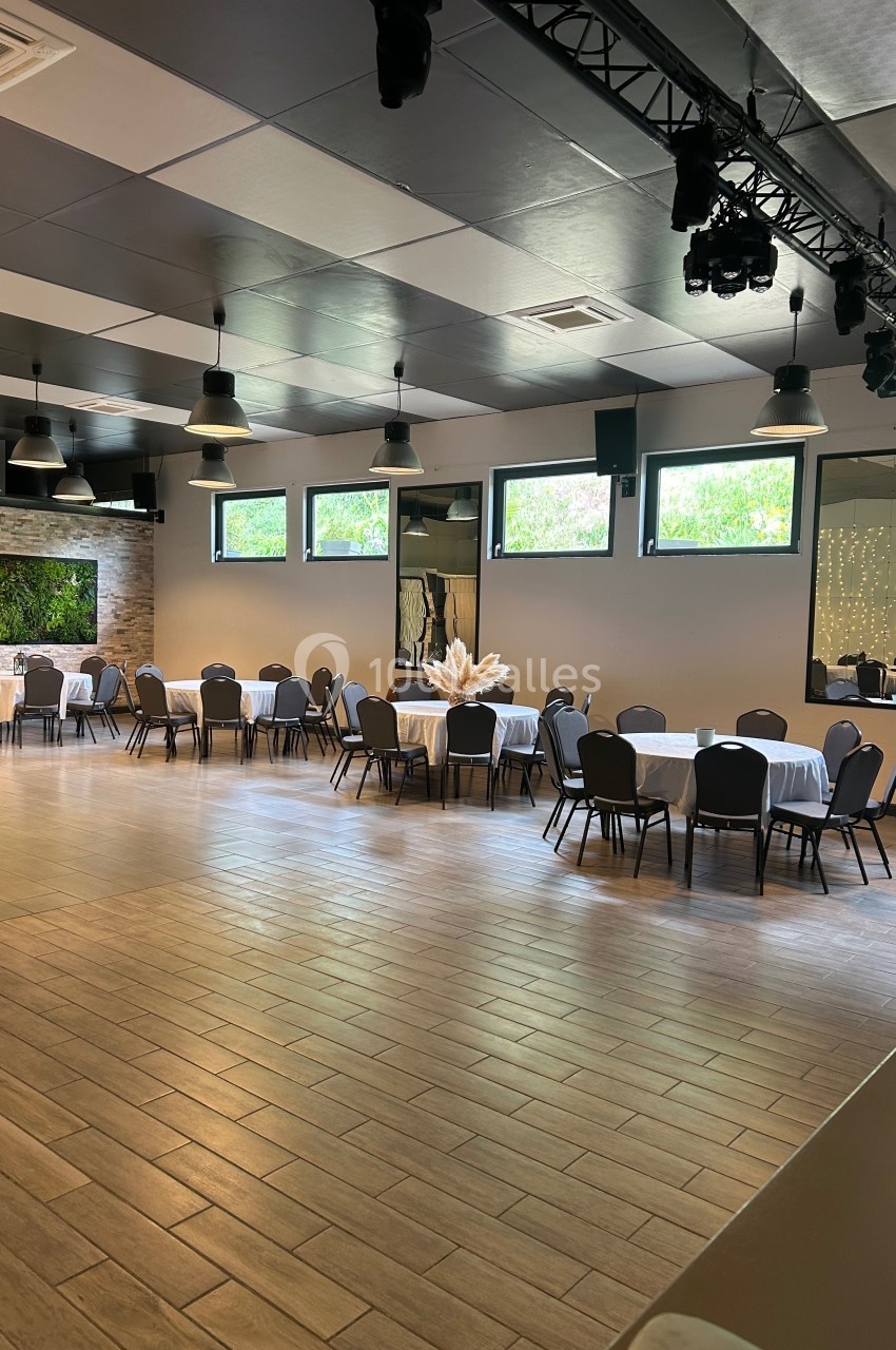 Salle de réception lumineuse avec tables rondes dressées, chaises noires et grandes fenêtres donnant sur l'extérieur.