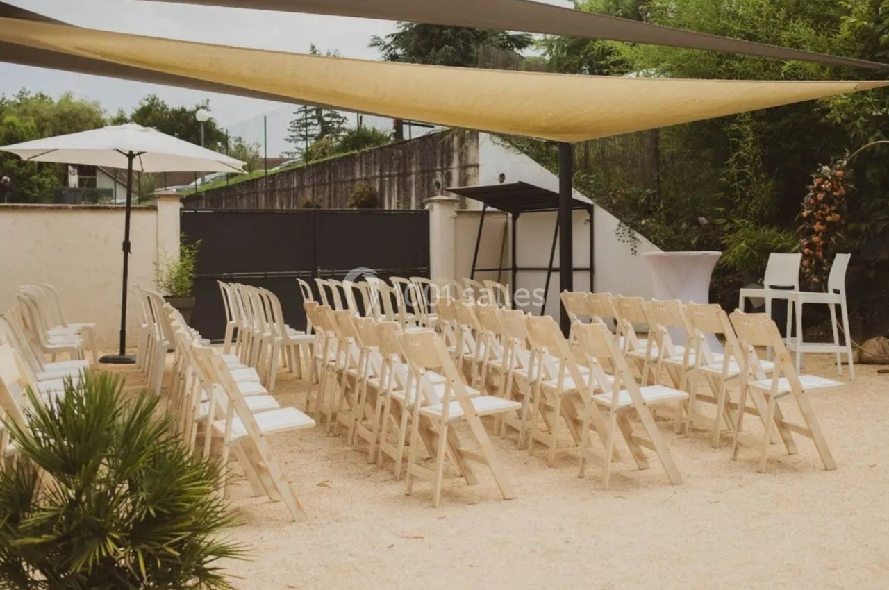 Chaises blanches alignées en extérieur sous des voiles d'ombrage, avec un pupitre et des plantes en arrière-plan.