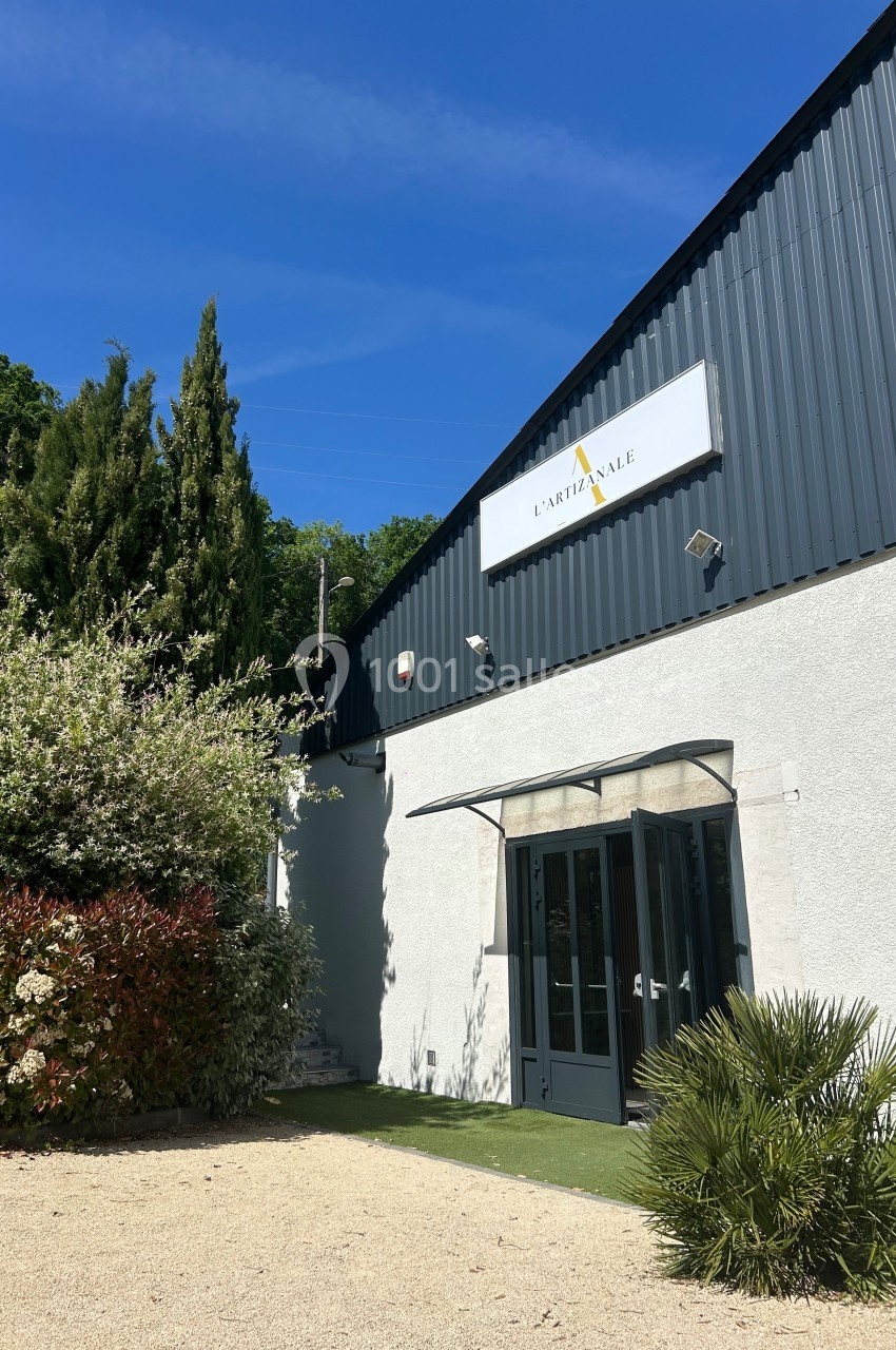 Façade d'un bâtiment moderne avec un panneau ’L'Artisanale’, entouré de végétation et éclairé par un ciel bleu.