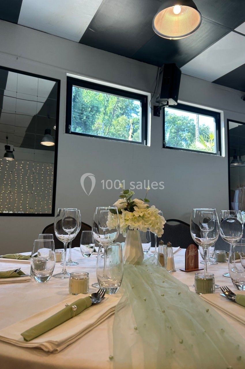 Table dressée avec nappes blanches, verres, couverts et centre de table floral, dans une salle lumineuse avec grandes…