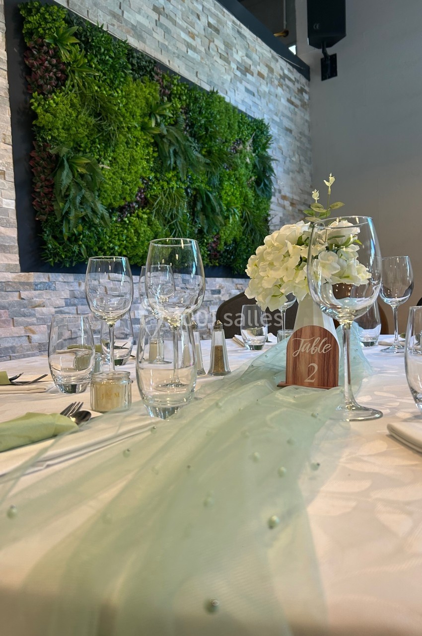 Table dressée avec nappe blanche, décoration en tissu vert, verres, fleurs blanches et mur végétal en arrière-plan.