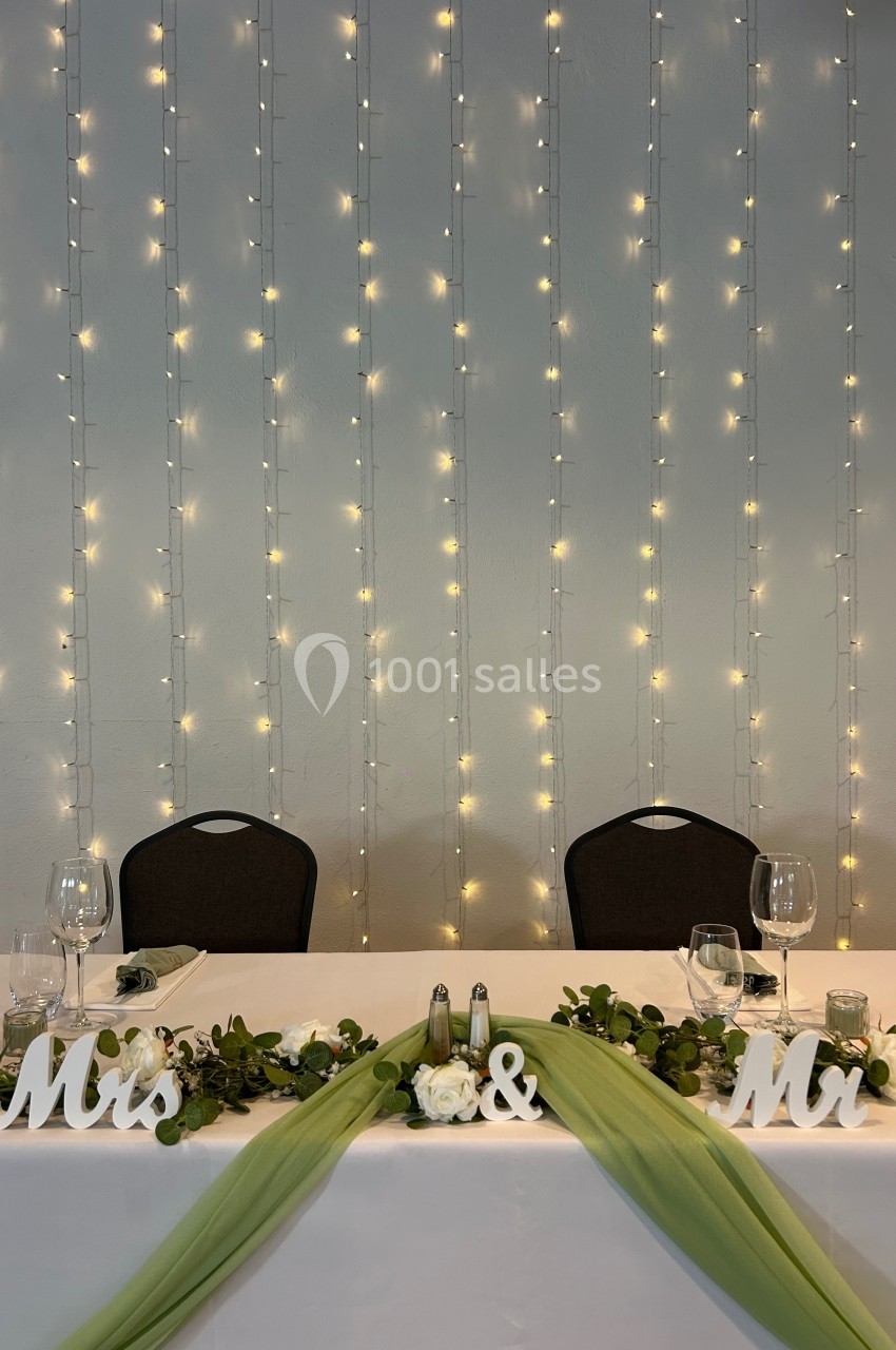 Table de mariage décorée avec des guirlandes lumineuses, des feuillages, des fleurs blanches et des lettres ’Mr & Mrs’.