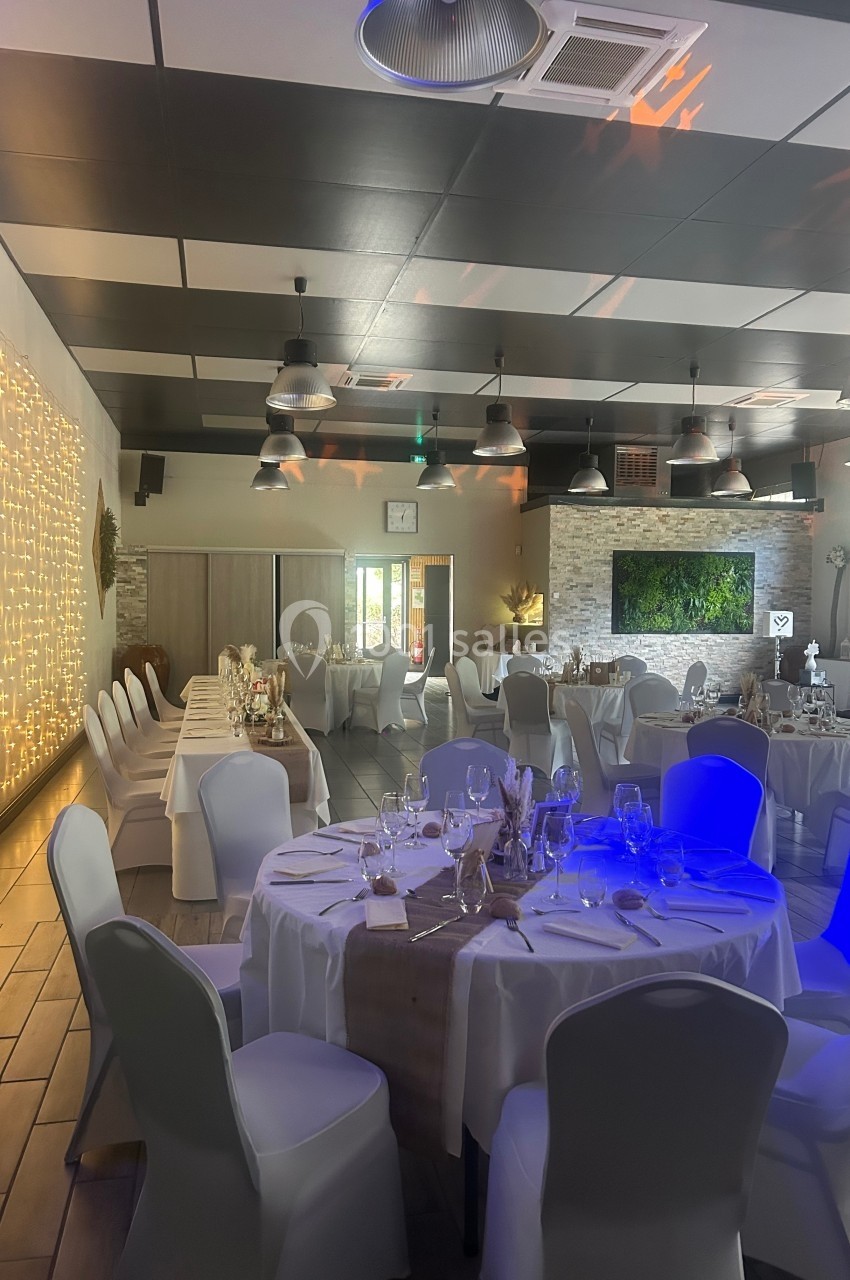 Salle de réception décorée avec des tables rondes dressées, des chaises blanches et un mur illuminé de guirlandes.
