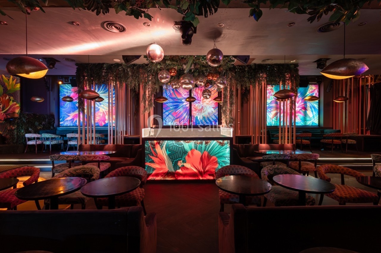 Salle de bar colorée avec décorations florales, éclairage tamisé et écrans affichant des motifs lumineux.