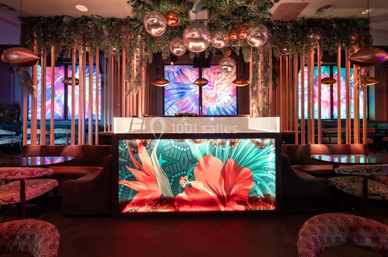 Bar avec comptoir lumineux décoré de motifs floraux, entouré de plantes suspendues et d'écrans colorés en arrière-plan.