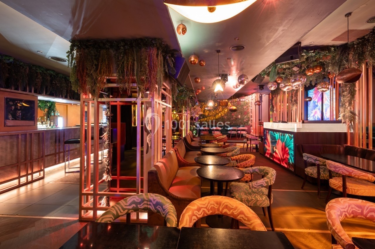 Intérieur d'un bar ou restaurant coloré avec des tables rondes, des chaises décorées et un éclairage tamisé.