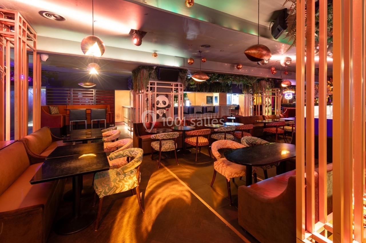 Salle de restaurant ou bar avec tables, chaises colorées, éclairage tamisé et décoration végétale suspendue.