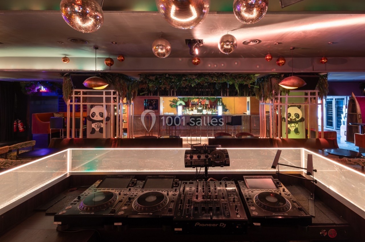 Poste de DJ avec platines en premier plan, espace lounge avec bar, décorations végétales et lumières suspendues en arrière…