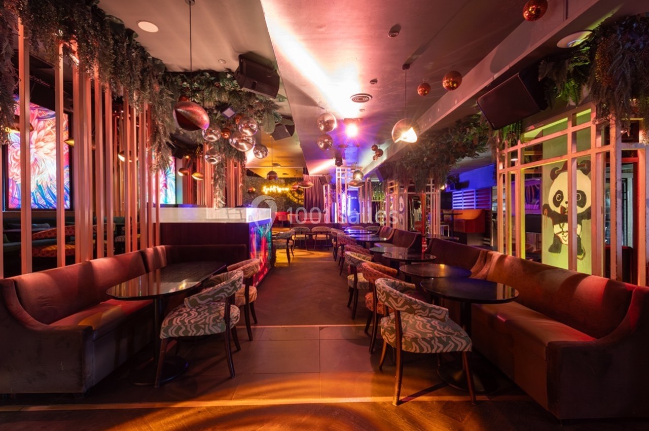 Salle de restaurant ou bar avec éclairage coloré, banquettes, tables rondes et décoration végétale et lumineuse.