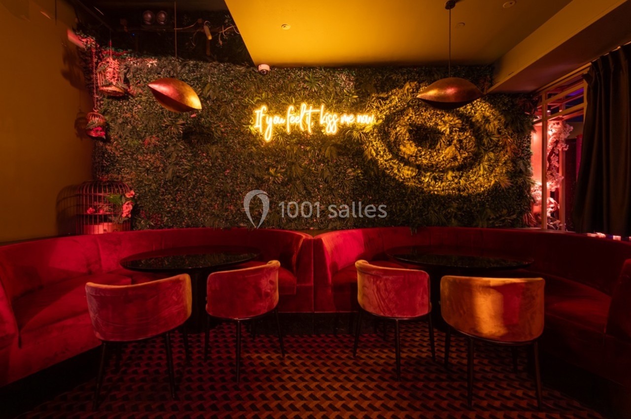 Salle avec banquettes rouges, tables rondes, mur végétal éclairé par un néon jaune affichant un message.