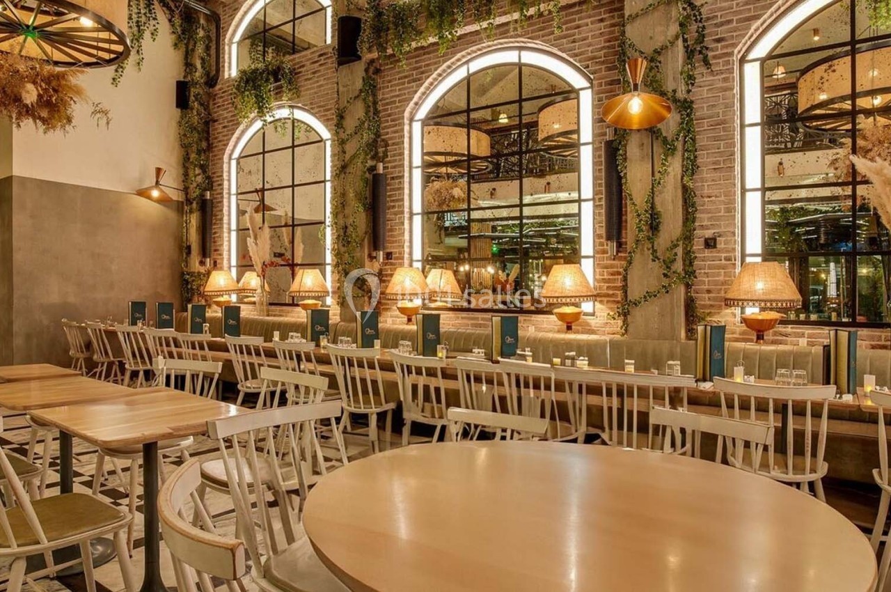 Salle de restaurant chaleureuse avec tables en bois clair, chaises blanches, miroirs et plantes suspendues.