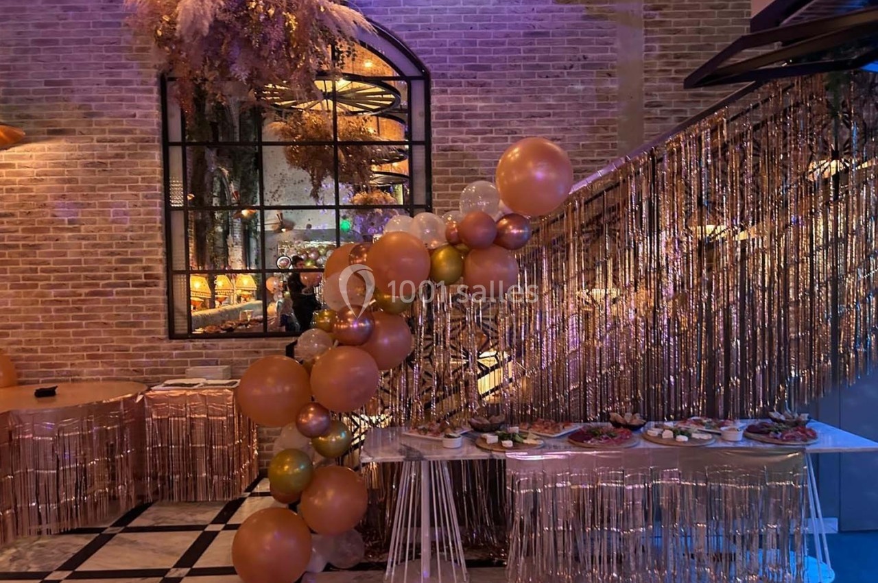 Décoration festive avec ballons dorés et roses, guirlandes scintillantes et table garnie de plats dans une salle en briques.