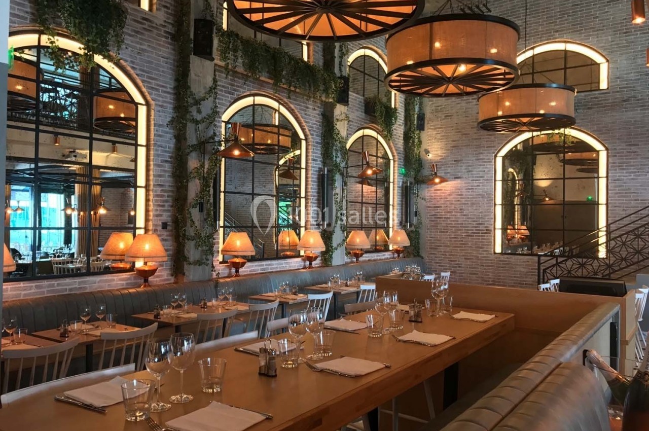 Salle de restaurant élégante avec tables dressées, grandes fenêtres en arc, murs en briques et luminaires suspendus.
