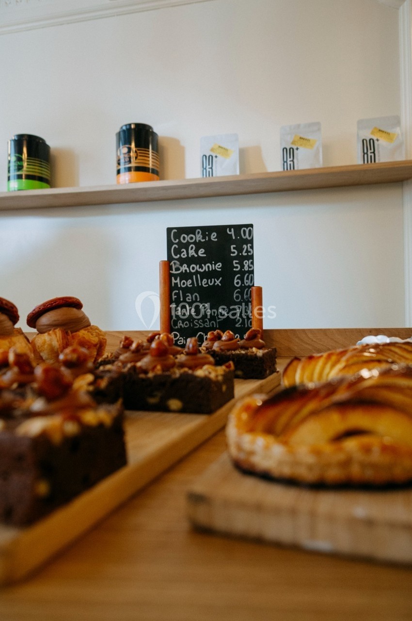 Pâtisseries variées disposées sur un comptoir en bois avec une ardoise indiquant les prix en arrière-plan.