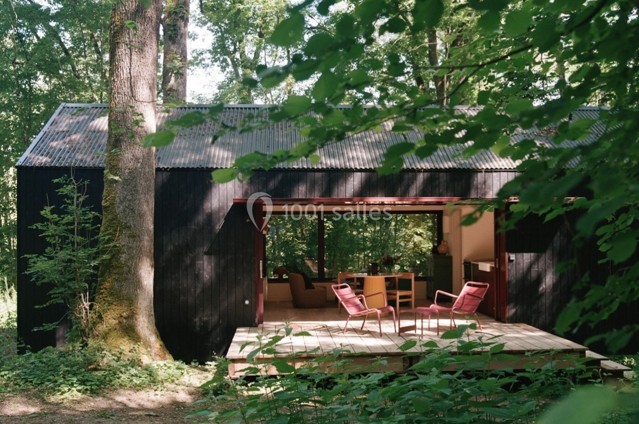 Cabane en bois sombre avec terrasse ouverte, entourée de forêt verdoyante et éclairée par la lumière naturelle.