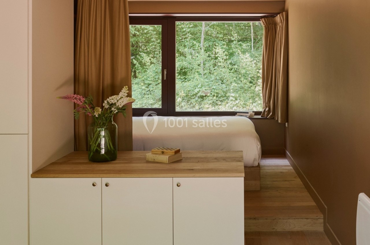 Chambre lumineuse avec lit, grandes fenêtres donnant sur une forêt, meuble en bois clair et vase de fleurs.