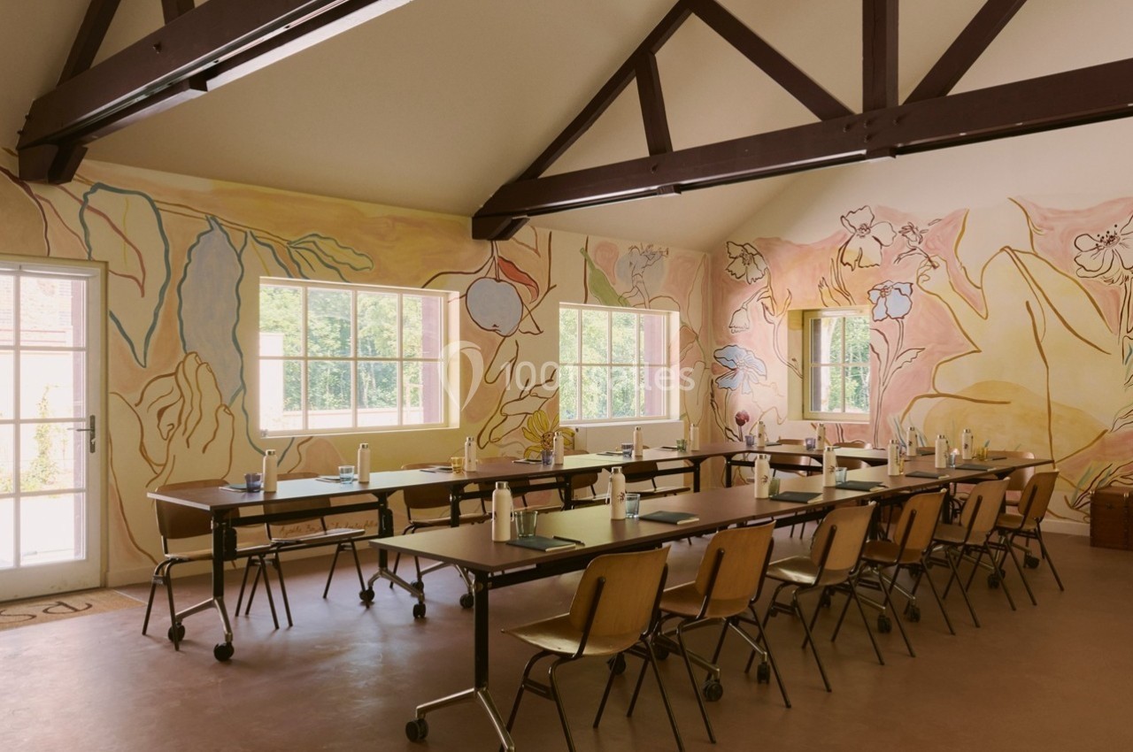 Salle lumineuse avec tables alignées, chaises en bois, murs décorés de fresques florales et grandes fenêtres.