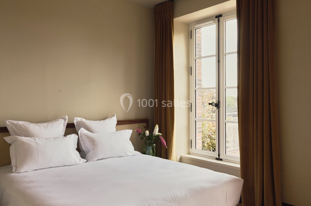 Lit double avec draps blancs dans une chambre lumineuse, fenêtre ouverte avec rideaux marron et vase de fleurs.
