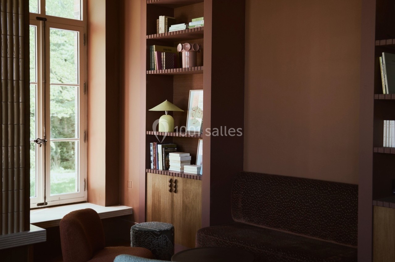 Salon chaleureux avec fauteuils, table basse, étagères remplies de livres et lampe, éclairé par une fenêtre.