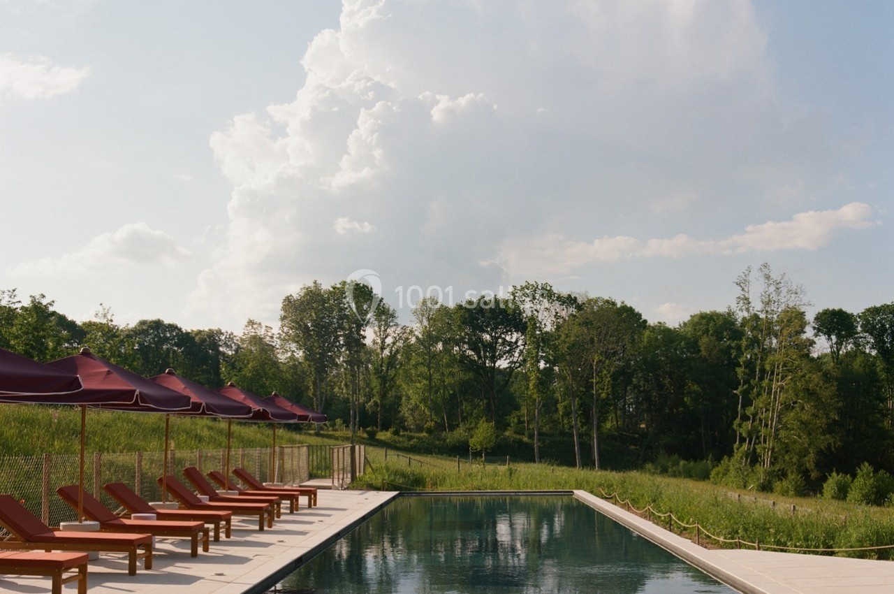 Piscine extérieure entourée de chaises longues et parasols rouges, avec vue sur un paysage verdoyant et un ciel…
