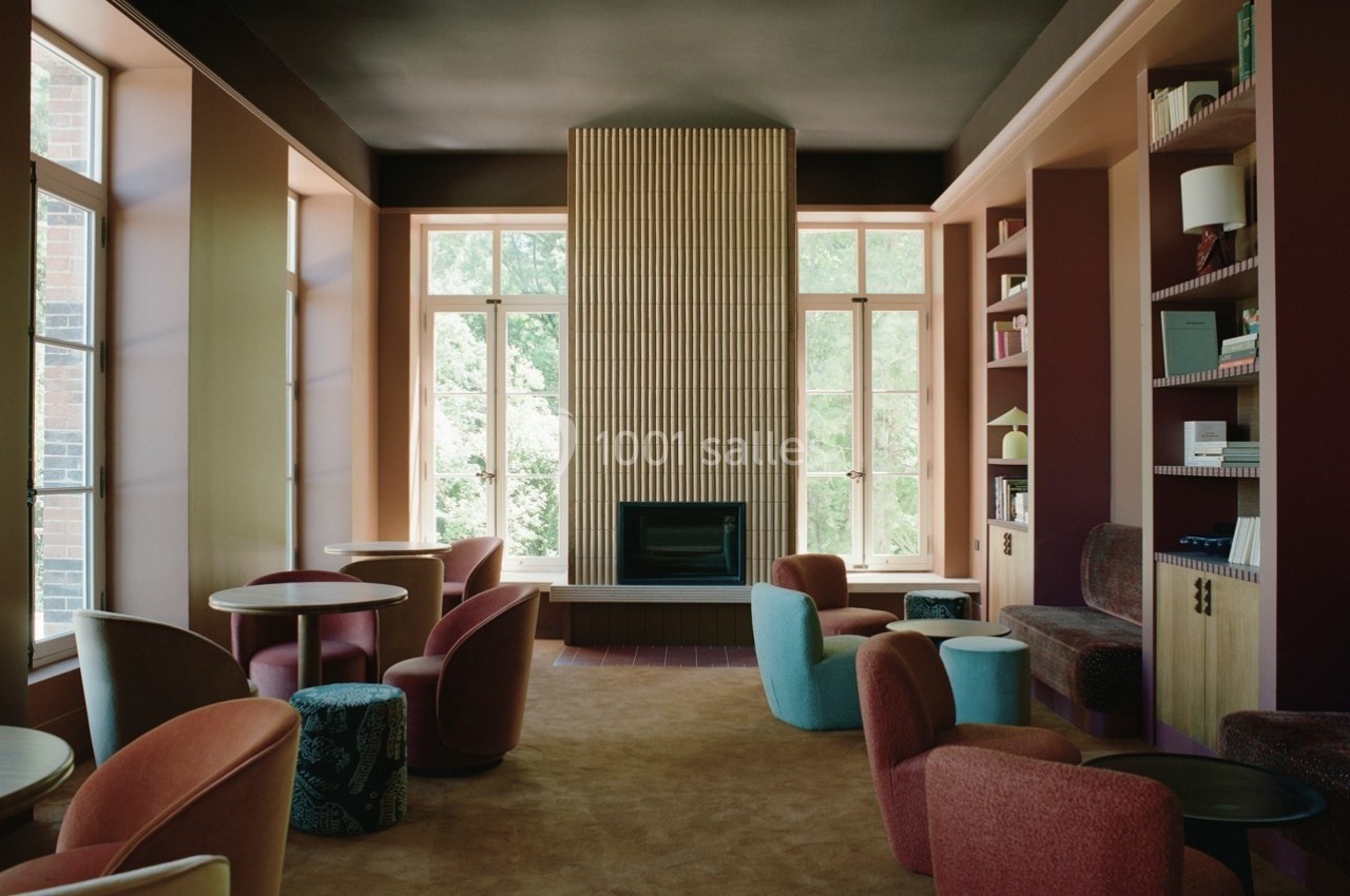 Salle de lecture lumineuse avec cheminée centrale, fauteuils colorés et étagères remplies de livres.