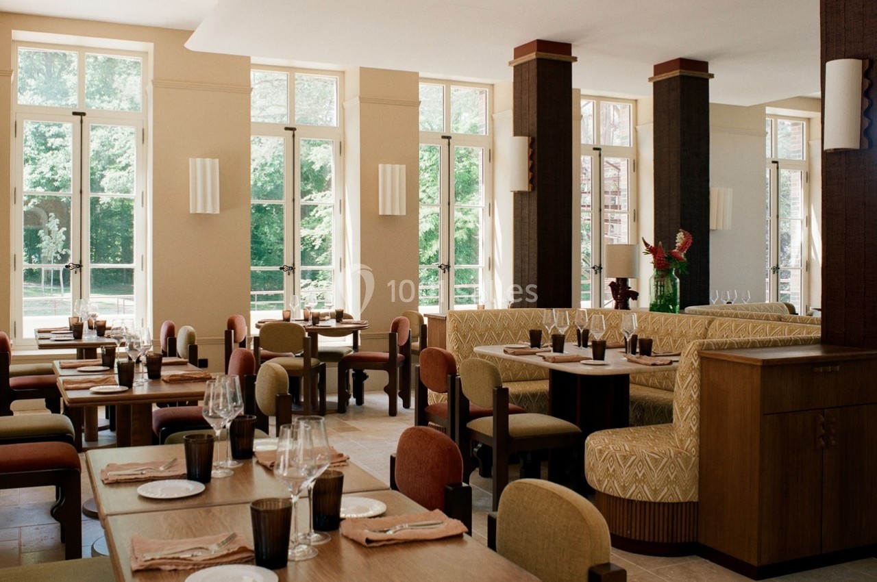 Salle de restaurant lumineuse avec grandes baies vitrées, tables dressées et banquettes beige.