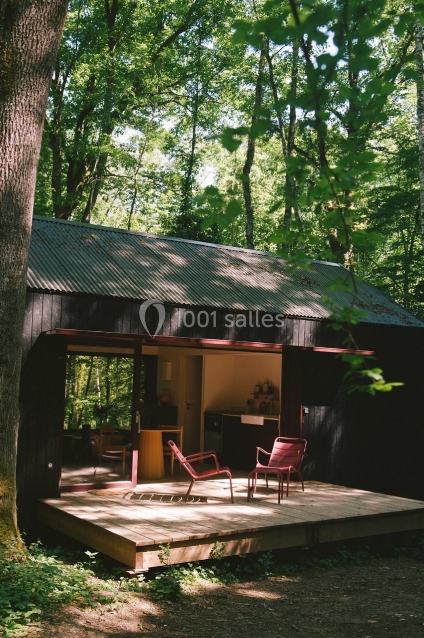 Petite maison en bois sombre avec terrasse, entourée d'arbres dans une forêt verdoyante.