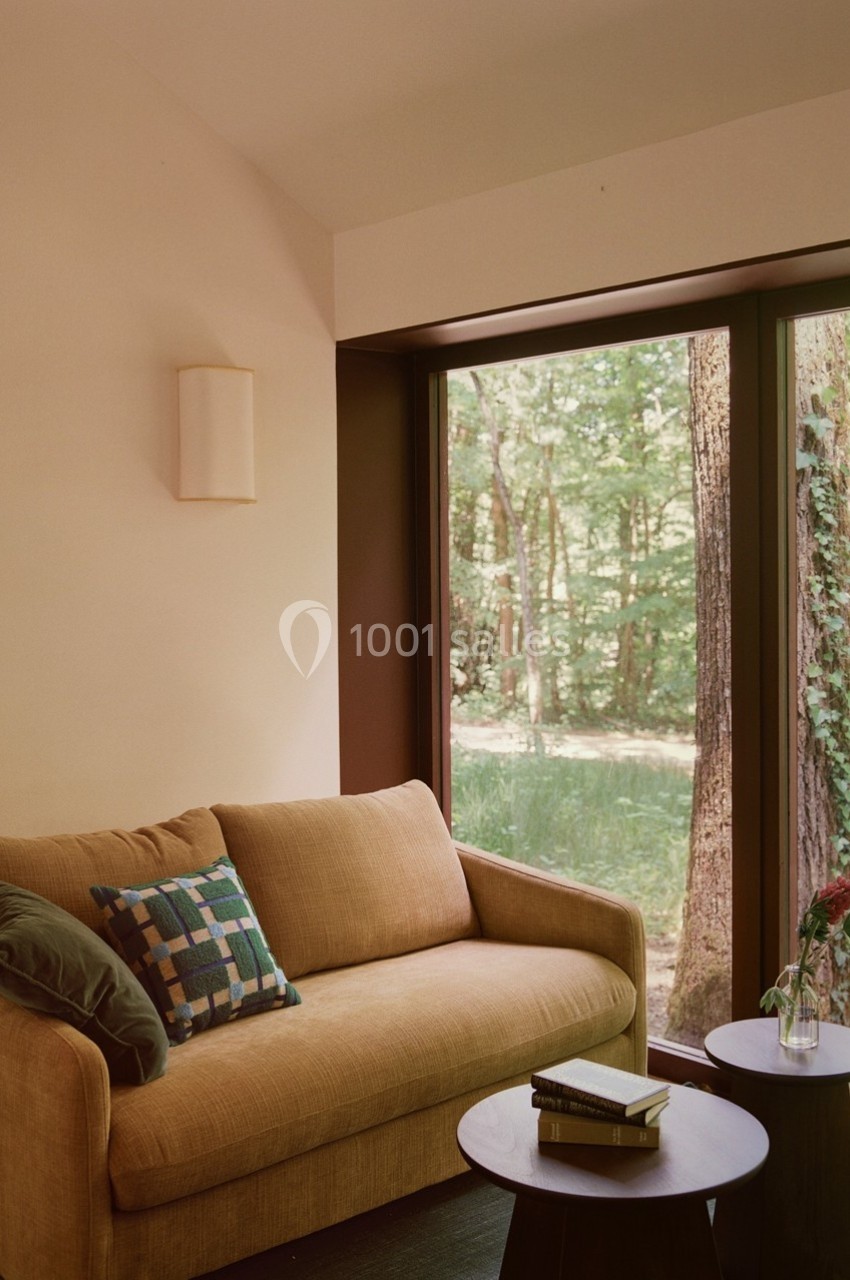 Canapé beige avec coussins, tables basses et grande fenêtre donnant sur une forêt verdoyante.
