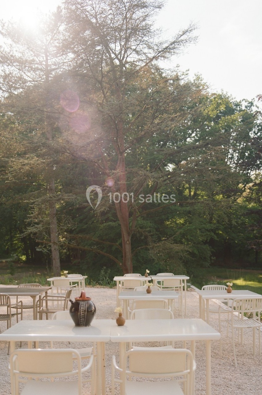 Tables et chaises blanches disposées en extérieur sur une terrasse en gravier, entourées d'arbres et de verdure.