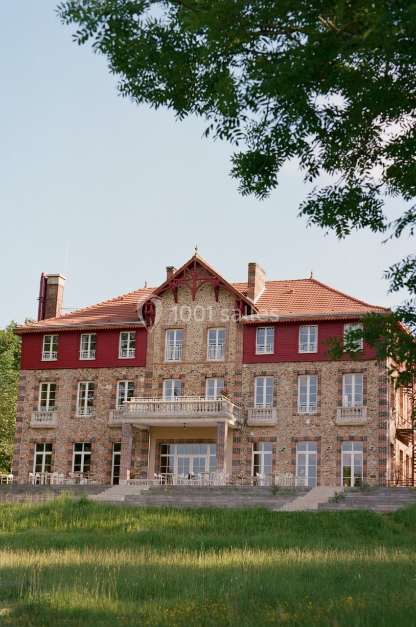 Façade d'un grand bâtiment en briques rouges et pierres, entouré de verdure sous un ciel dégagé.
