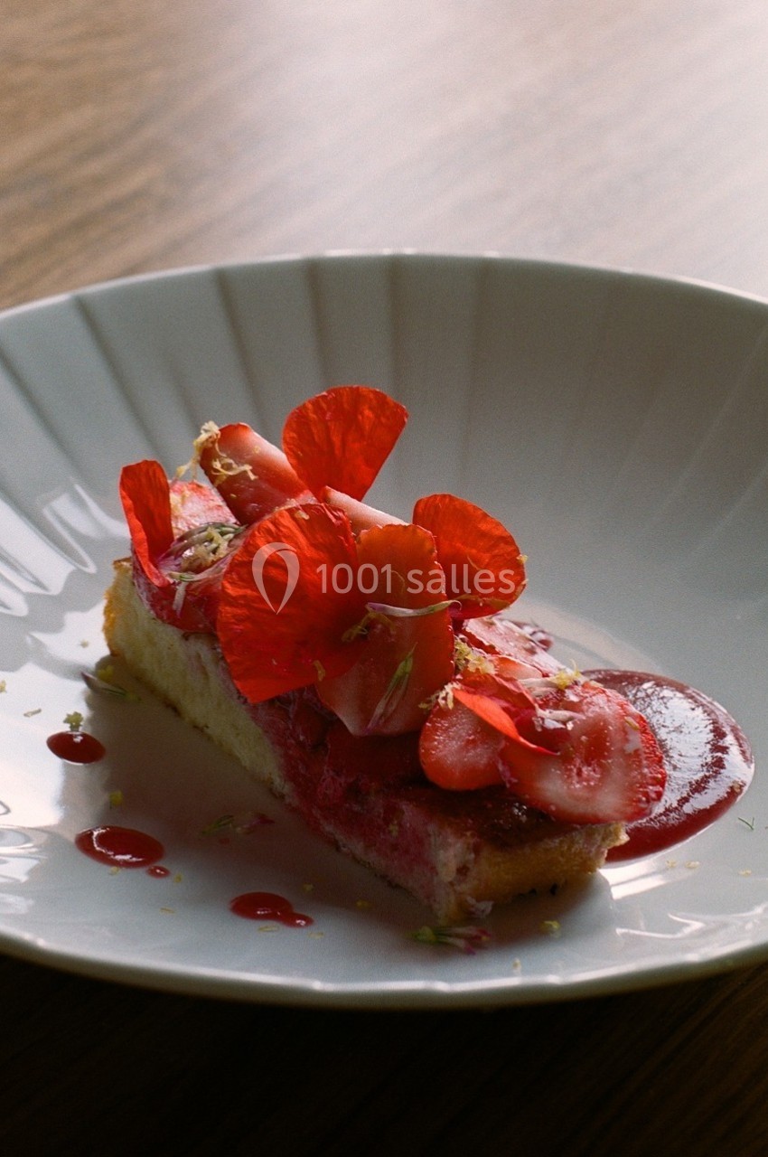 Part de gâteau garnie de fraises, pétales rouges et sauce, servie dans une assiette blanche sur une table en bois.