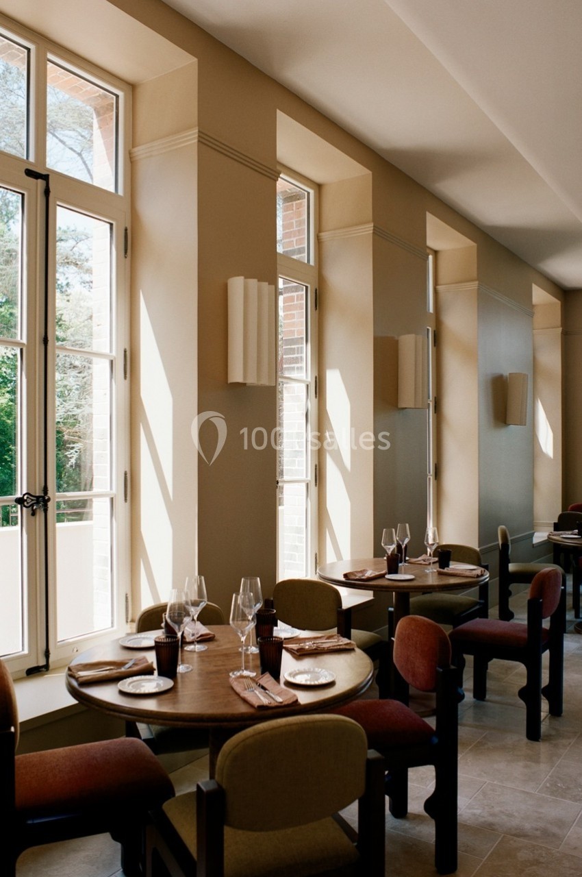 Salle de restaurant lumineuse avec de grandes fenêtres, des tables rondes dressées et des chaises rembourrées.