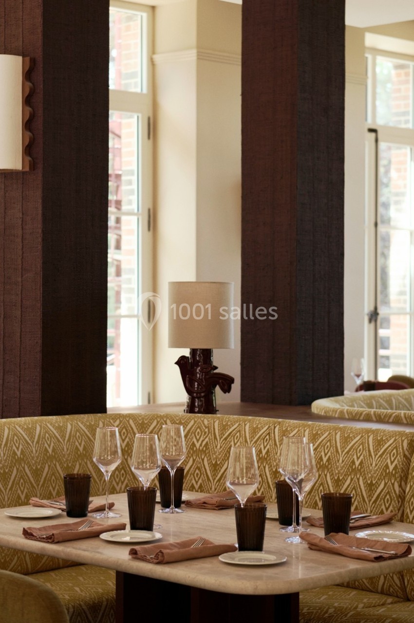 Banquette beige avec table dressée, verres et serviettes, dans un restaurant lumineux avec grandes fenêtres.