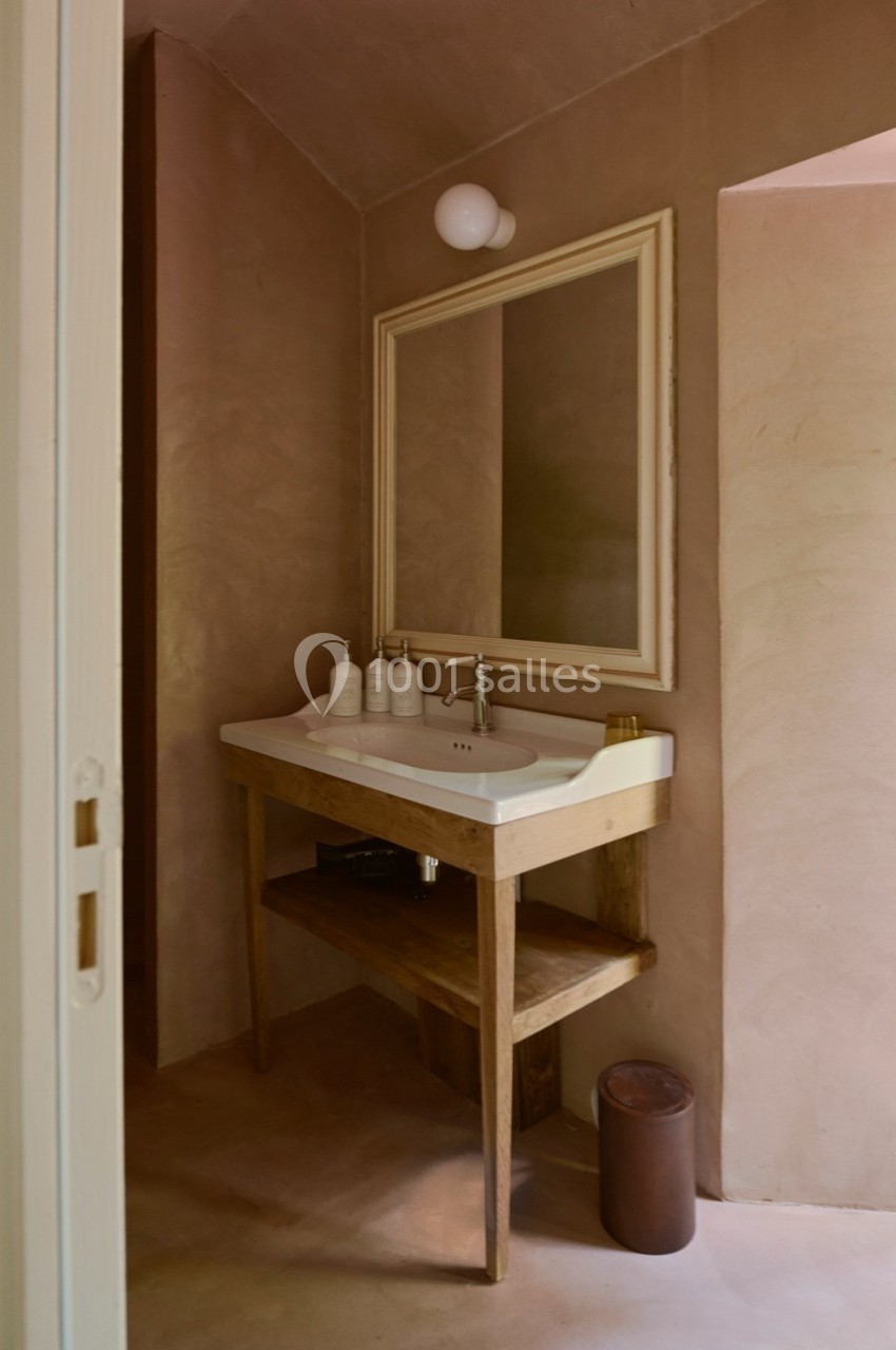 Lavabo en céramique blanche avec miroir encadré, étagère en bois et accessoires dans une salle de bain aux tons beige.