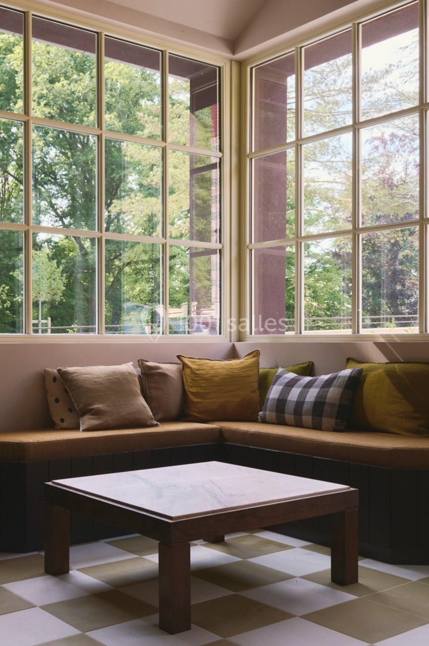 Coin salon lumineux avec banquette en bois, coussins colorés et table basse, donnant sur un jardin verdoyant.