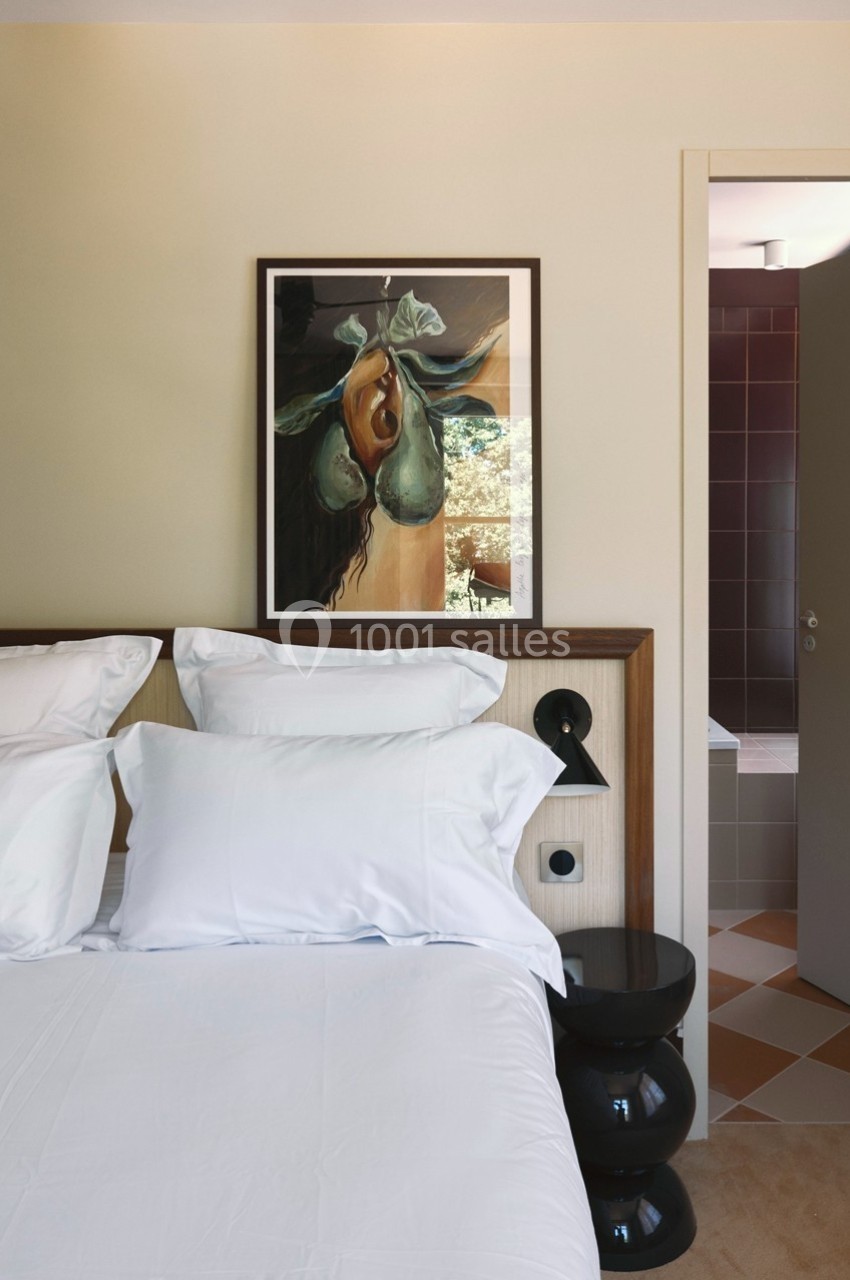 Lit avec draps blancs dans une chambre lumineuse, tableau encadré au mur et porte ouverte sur une salle de bain.