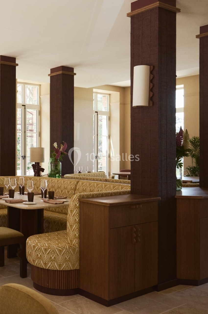 Salle de restaurant élégante avec banquettes en tissu à motifs, colonnes en bois sombre et grandes fenêtres lumineuses.