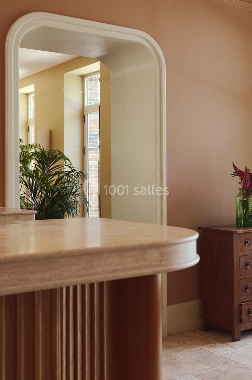 Miroir encadré blanc reflétant une pièce lumineuse avec plantes, meuble en bois et vase de fleurs sur une commode.