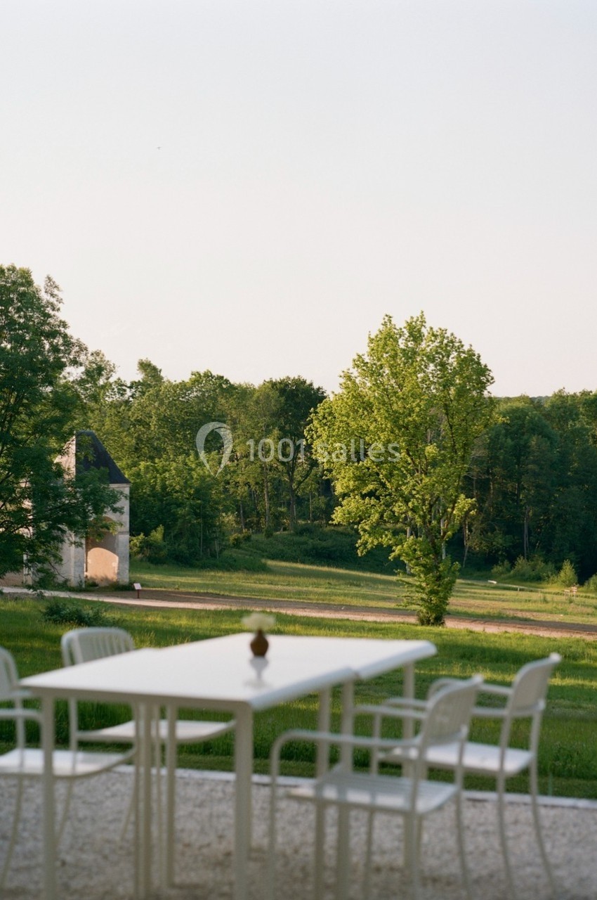 Table et chaises blanches sur une terrasse avec vue sur un paysage verdoyant et une petite bâtisse au loin.