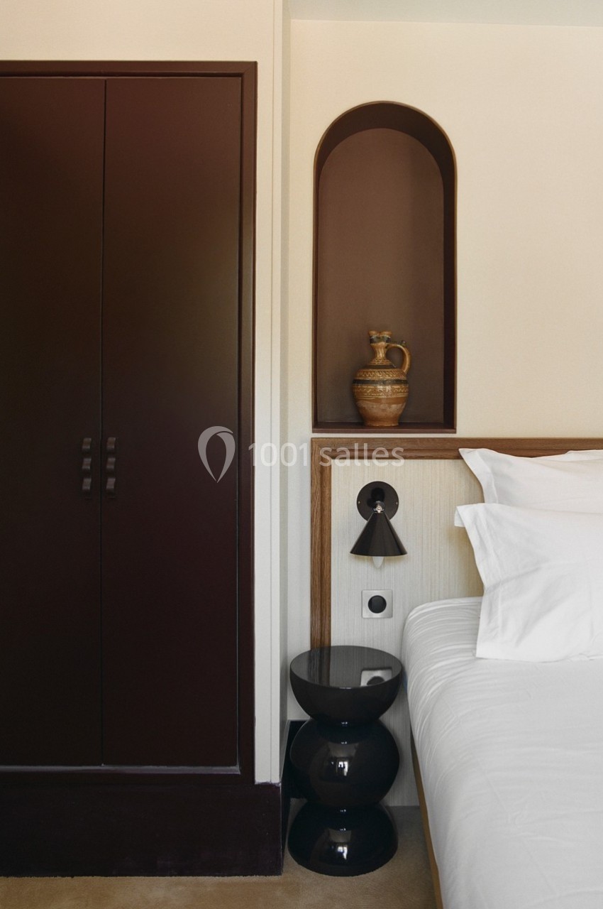 Placard en bois sombre, niche murale avec une cruche en céramique, table de chevet noire et lit avec oreillers blancs.