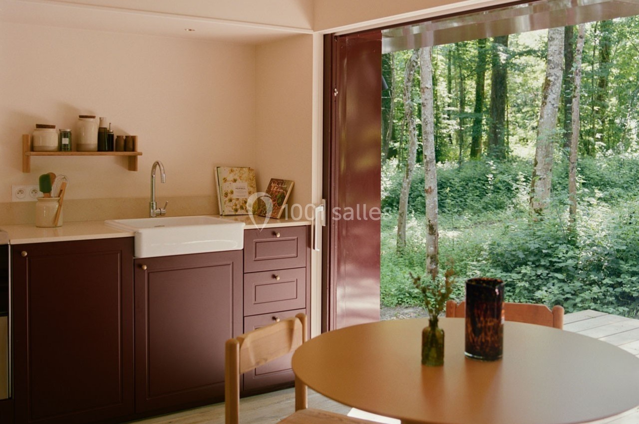 Cuisine ouverte avec meubles bordeaux, table ronde en bois et vue sur une forêt à travers une grande baie vitrée.