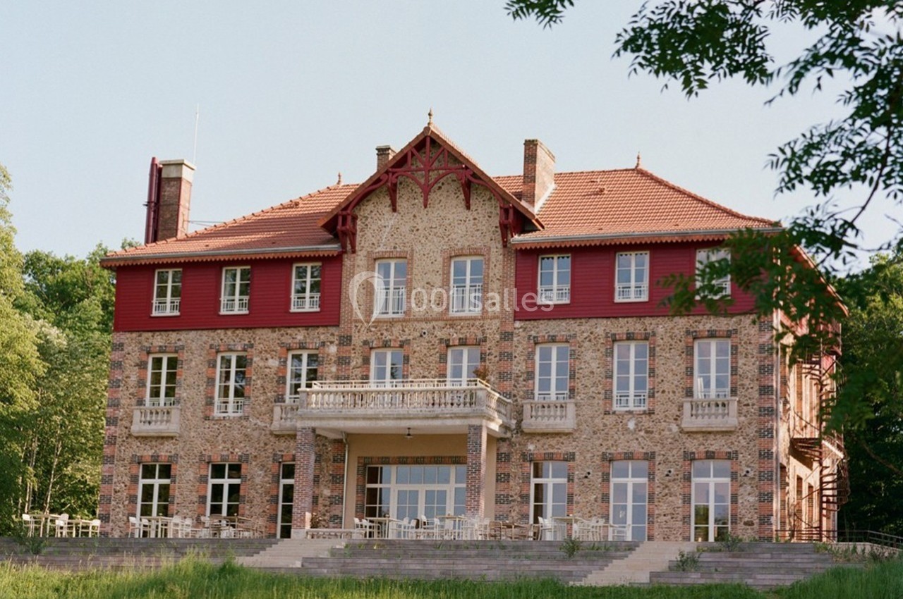 Façade d'un grand bâtiment en briques avec des détails rouges, entouré de verdure et éclairé par la lumière naturelle.