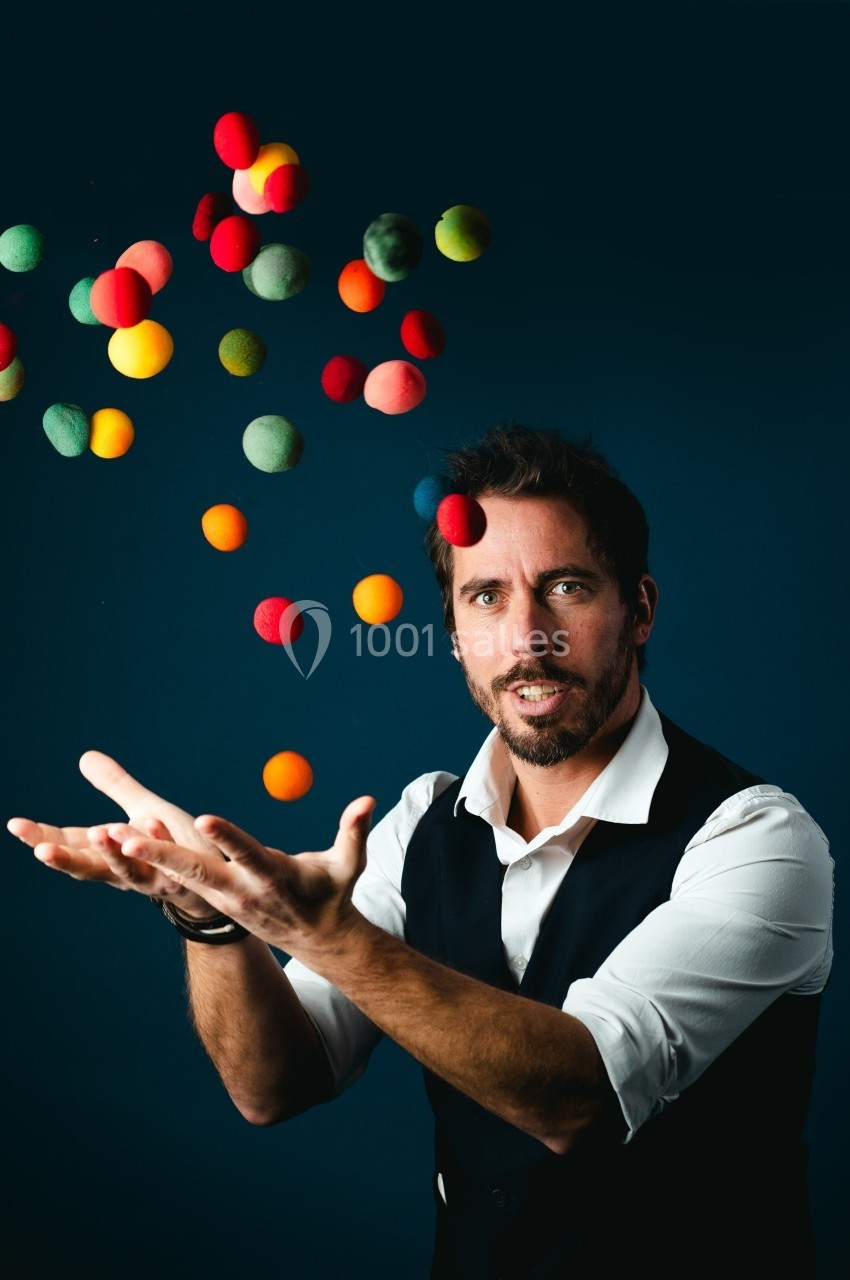 Un homme en chemise et gilet lance des balles colorées en l'air sur un fond sombre.