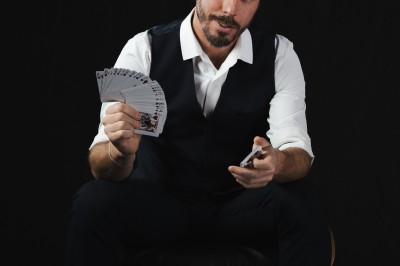 Un homme assis en costume décontracté, tenant un éventail de cartes dans une main, sur fond noir.