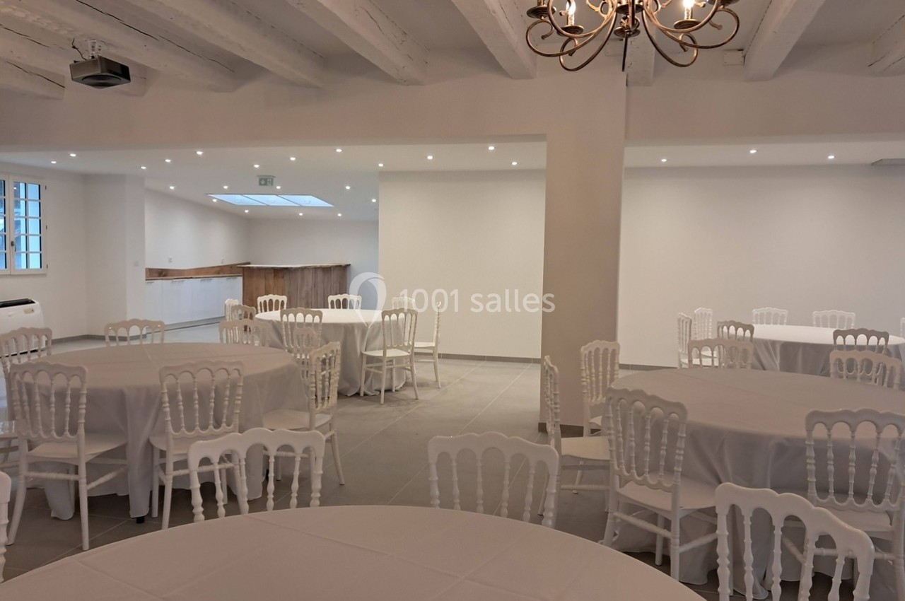 Salle lumineuse avec tables rondes dressées de nappes blanches, chaises blanches et plafond aux poutres apparentes.