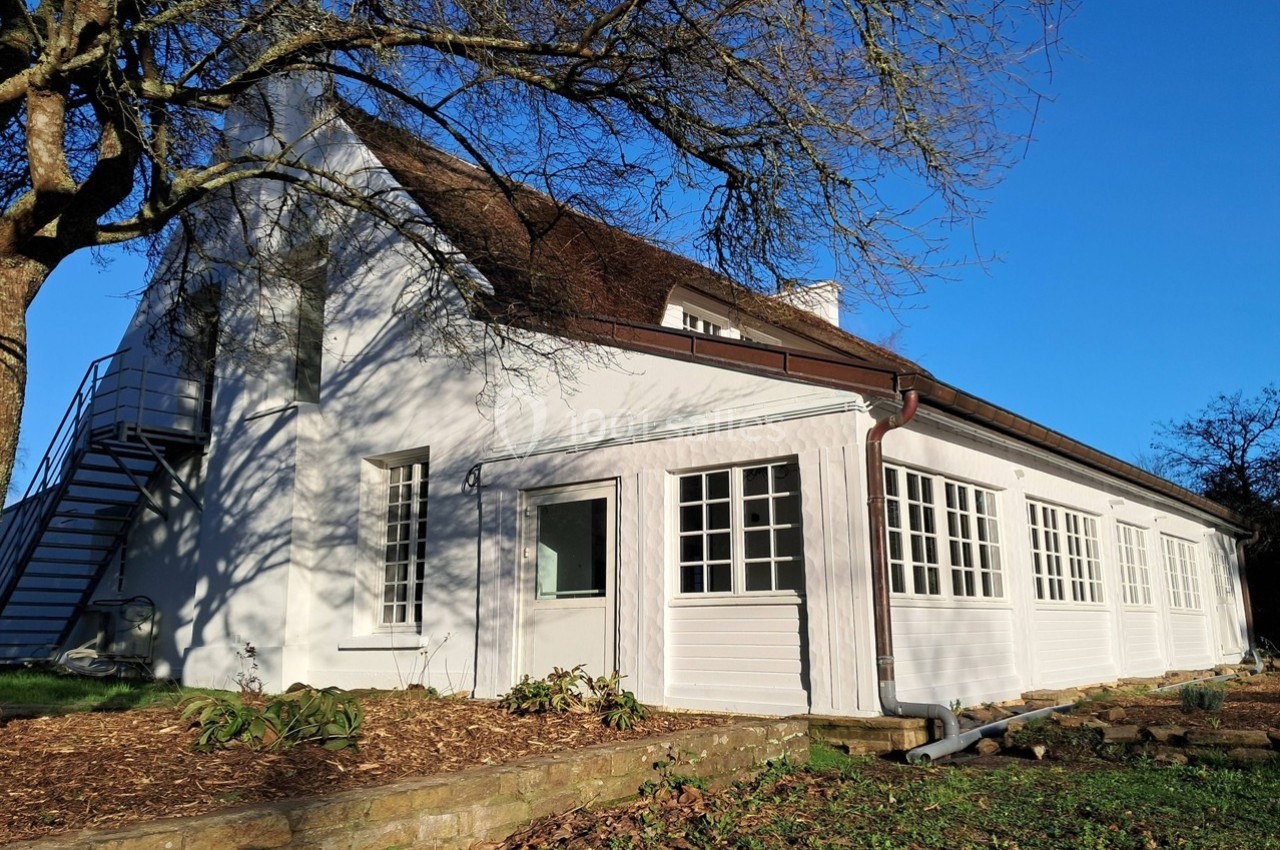 Maison blanche avec toit en pente, entourée d'arbres, sous un ciel bleu clair.